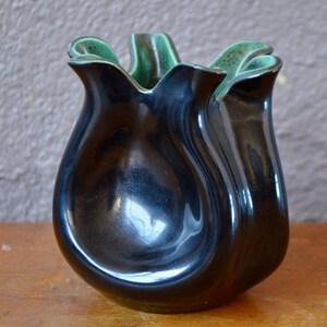 May include: A glossy black ceramic vase with a unique, tulip-like shape. The rim of the vase has a ruffled edge with a contrasting green interior. The vase sits on a wooden surface, showcasing its elegant design.