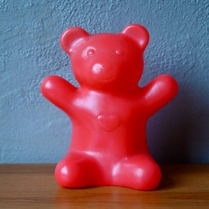May include: A bright red plastic teddy bear figurine with outstretched arms and a heart-shaped detail on its chest. The toy is sitting on a wooden surface against a blue wall.