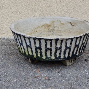 May include: A large, round, concrete planter with a black and white geometric pattern. The planter has three short legs and is sitting on a grey surface.