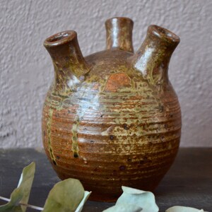 Peut inclure: Un vase en céramique brun avec trois ouvertures, présentant un aspect rustique, fait à la main, avec un glaçage marbré.