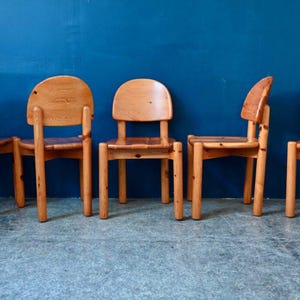 May include: Five wooden chairs with a natural wood finish. The chairs have a simple design with a rounded back and four legs. The chairs are arranged in a row against a blue wall.