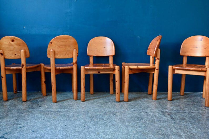 May include: Five wooden chairs with a natural wood finish. The chairs have a simple design with a rounded back and four legs. The chairs are arranged in a row against a blue wall.