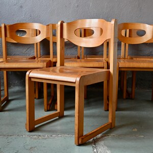 May include: Set of six wooden chairs with a mid-century modern design. The chairs have a light brown finish and feature a unique cutout in the back.