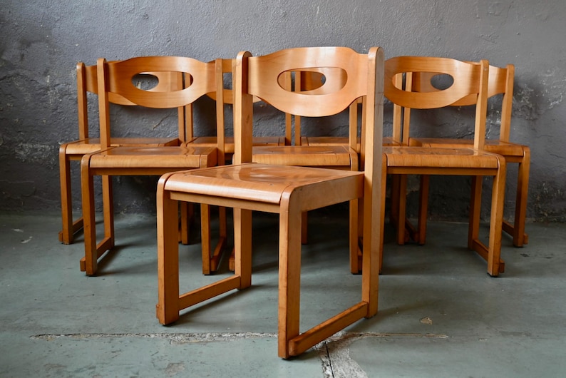 May include: Set of six wooden chairs with a mid-century modern design. The chairs have a light brown finish and feature a unique cutout in the back.