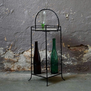 Puede incluir: Un estante de vino de metal negro con dos botellas de vidrio, una verde y una marrón, y un vaso en el estante superior. El estante está de pie sobre un suelo de hormigón frente a una pared gris texturizada.