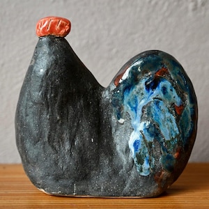 Peut inclure: Une figurine de coq en céramique avec un corps noir mat à la surface texturée. La tête est surmontée d'une crête orange vif. La partie de la queue présente un vernis brillant et tacheté dans des tons de bleu profond, blanc et brun contrastés.