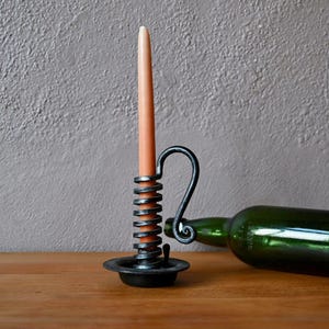 Puede incluir: Un portavelas de metal hecho a mano con un diseño en espiral, que sostiene una vela cónica de color melocotón. Una botella de vidrio verde descansa sobre una superficie de madera, junto al portavelas. El fondo es una pared gris texturizada.