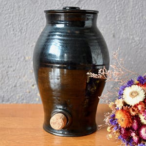 May include: A black ceramic jar with a cork stopper. The jar has a wide mouth and a rounded body. It is sitting on a wooden surface.