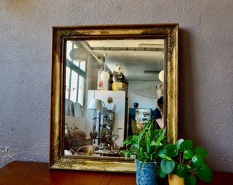 May include: An ornate mirror with a gold-toned distressed finish. The mirror is reflecting a room with a white ceiling and a person standing in front of it.
