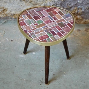Peut inclure: Petit support de plante à trois pieds avec un dessus en mosaïque. Le dessus présente un motif de carreaux rectangulaires dans les tons de rouge, marron et blanc, encadré d'un bord en métal doré. Les pieds sont marron foncé.