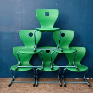 Peut inclure: Six chaises vertes empilées, avec un design distinctif et un trou circulaire dans le dossier. Les chaises sont posées contre un mur bleu foncé. Les pieds sont en métal bleu clair.
