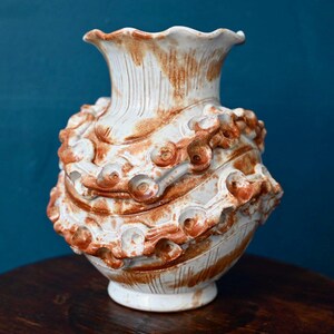 Peut inclure: Un vase en céramique avec une base blanche et des accents de glaçure brune striée. Le vase présente un bord ondulé et un corps large et arrondi, orné de deux bandes horizontales saillantes de motifs en relief circulaires et floraux.