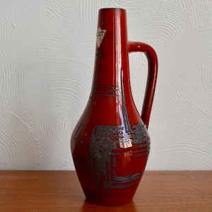 May include: A red ceramic vase with a geometric design in grey and purple. The vase has a handle and a narrow neck.