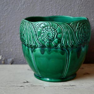 May include: A green ceramic planter with an ornate floral design. The planter has a wide, rounded base and a slightly flared rim.