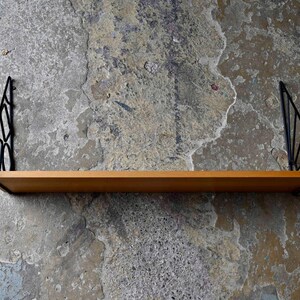 May include: A wooden shelf with black metal brackets. The shelf is rectangular and made of light-colored wood. The brackets are triangular with a geometric design. The shelf is mounted on a textured, gray wall.