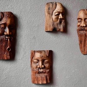 Peut inclure: Quatre sculptures en bois de visages avec des barbes, chacune avec une expression unique. Les sculptures sont montées sur un mur gris clair.