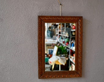 May include: A wooden framed mirror with an ornate design. The mirror reflects a cluttered room with shelves filled with various items.