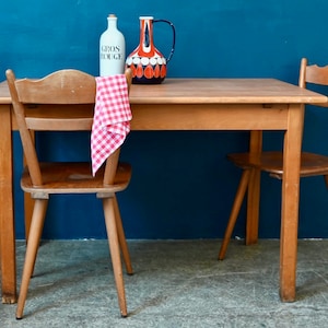 Peut inclure: Une table en bois avec deux chaises. La table est recouverte d'une nappe à carreaux rouge et blanc qui est drapé sur l'une des chaises. Une bouteille blanche avec du texte noir qui lit "Gros Rouge" et un pichet en céramique coloré sont sur la table.