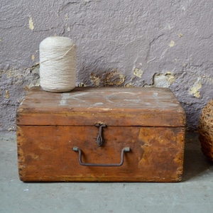 May include: A wooden box with a metal handle and a latch. The box is sitting on a concrete floor in front of a textured purple wall. A ball of twine and a wicker-wrapped bottle are also in the image.