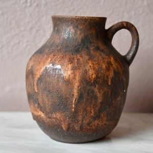 Puede incluir: Un jarrón de cerámica marrón con una superficie texturizada y una sola asa. El jarrón tiene un cuerpo redondeado y una abertura ligeramente acampanada.