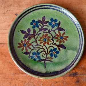 May include: A green ceramic plate with a hand-painted floral design. The flowers are blue, orange, and brown with green leaves. The plate has a dark blue rim.