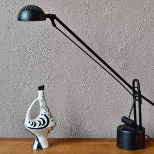 May include: A black adjustable desk lamp with a round shade and a long, articulated arm. The lamp is on a wooden surface next to a white ceramic vase with a black and white design.