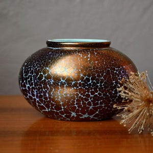 Peut inclure: Un vase en céramique arrondi et trapu présentant une finition bronze métallique avec un motif craquelé blanc distinct sur toute sa surface. L'intérieur de l'ouverture du vase est de couleur turquoise clair et le récipient a une texture lisse.