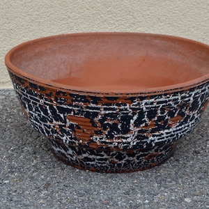 May include: A terracotta planter with a distressed black and white paint finish. The planter has a wide, round opening and a slightly flared base.