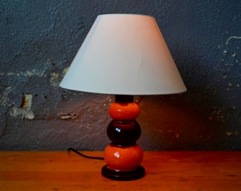 May include: A ceramic table lamp with a white lampshade. The base is made of three stacked spheres in shades of orange and brown.