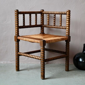 Può includere: Una sedia ad angolo in legno marrone scuro con una struttura tornita a rocchetto caratterizzata da dettagli sferici. La seduta è intrecciata in paglia naturale color beige chiaro e lo schienale è composto da due sezioni perpendicolari.