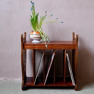 Peut inclure: Un meuble de rangement pour disques en bois foncé, contenant plusieurs vinyles. Un petit pot en céramique à rayures et une plante à fleurs bleues sont posés dessus. Le meuble a un design classique et vintage.