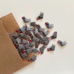 Könnte beinhalten: Ein brauner Papierumschlag, der mit grauen Origami-Vögeln überquillt. Die Vögel haben rote Schnäbel und sind in einem zufälligen Muster angeordnet.