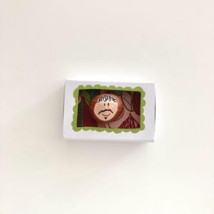 Könnte beinhalten: Eine kleine, handbemalte Holzfigur mit Gesicht, Schnurrbart und Haaren, präsentiert in einer weißen Schachtel mit grünem Rand. Der Hintergrund zeigt ein Blumenmuster mit roten und braunen Blättern.
