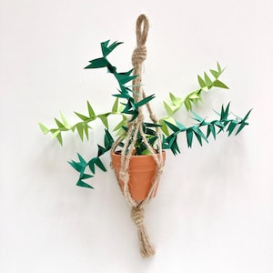 Könnte beinhalten: Ein handgefertigter Hängepflanzentopf mit einem Terrakotta-Topf und einem Makramee-Aufhänger aus Jute. Der Topf enthält eine Papierpflanze mit grünen und hellgrünen Blättern. Der Pflanzentopf hängt vor einem weißen Hintergrund.