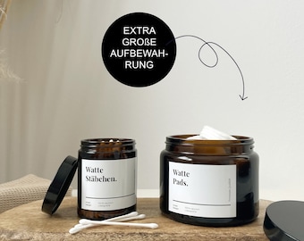 Puede incluir: Dos tarros de vidrio ámbar con tapas negras, uno lleno de bastoncillos de algodón y el otro con discos de algodón. Los tarros tienen etiquetas blancas con el texto "Watte Stäbchen." y "Watte Pads." en negro. Los tarros están sobre una superficie de madera.