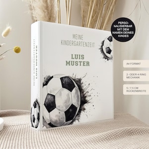 Könnte beinhalten: Ein weißer Ordner mit einem Fußball-Design und dem Text "MEINE KINDERGARTENZEIT LUIS MUSTER". Der Ordner hat auch den Text "A4 FORMAT", "2- ODER 4-RING MECHANIK" und "5/7,5 CM RÜCKENBREITE".