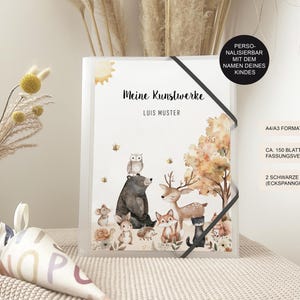 Könnte beinhalten: Ein weißer Ordner mit einer Aquarell-Illustration von Waldtieren, darunter ein Reh, ein Eule, ein Bär und ein Fuchs. Der Ordner hat den Text "Meine Kunstwerke" und "LUIS MUSTER" auf der Vorderseite. Der Ordner ist mit dem Namen eines Kindes personalisiert.