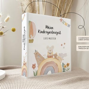 May include: A white binder with a colourful illustration of a hot air balloon, a rainbow, and animals. The text "Meine Kindergartenzeit" and "LUIS MUSTER" is printed on the front of the binder.