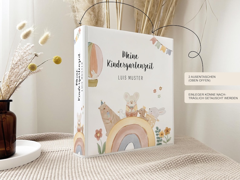 May include: A white binder with a colourful illustration of a hot air balloon, a rainbow, and animals. The text "Meine Kindergartenzeit" and "LUIS MUSTER" is printed on the front of the binder.