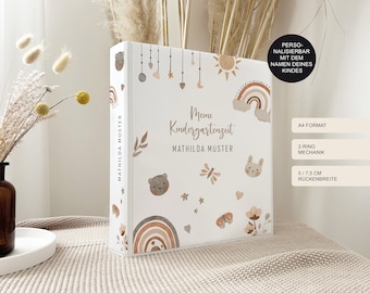 Puede incluir: Una carpeta A4 blanca con mecanismo de 2 anillas y un ancho de lomo de 5 o 7,5 cm. La cubierta presenta ilustraciones en marrón y beige de arcoíris, estrellas, un oso, un conejo y una mariposa, con el texto Meine Kindergartenzeit y Mathilda Muster.