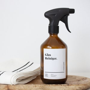 Könnte beinhalten: Eine braune Glasflasche mit schwarzer Sprühdüse, gefüllt mit Glasreiniger. Das Etikett lautet "Glas Reiniger.". Die Flasche steht auf einer Holzoberfläche mit einem weiß-schwarz gestreiften Handtuch.