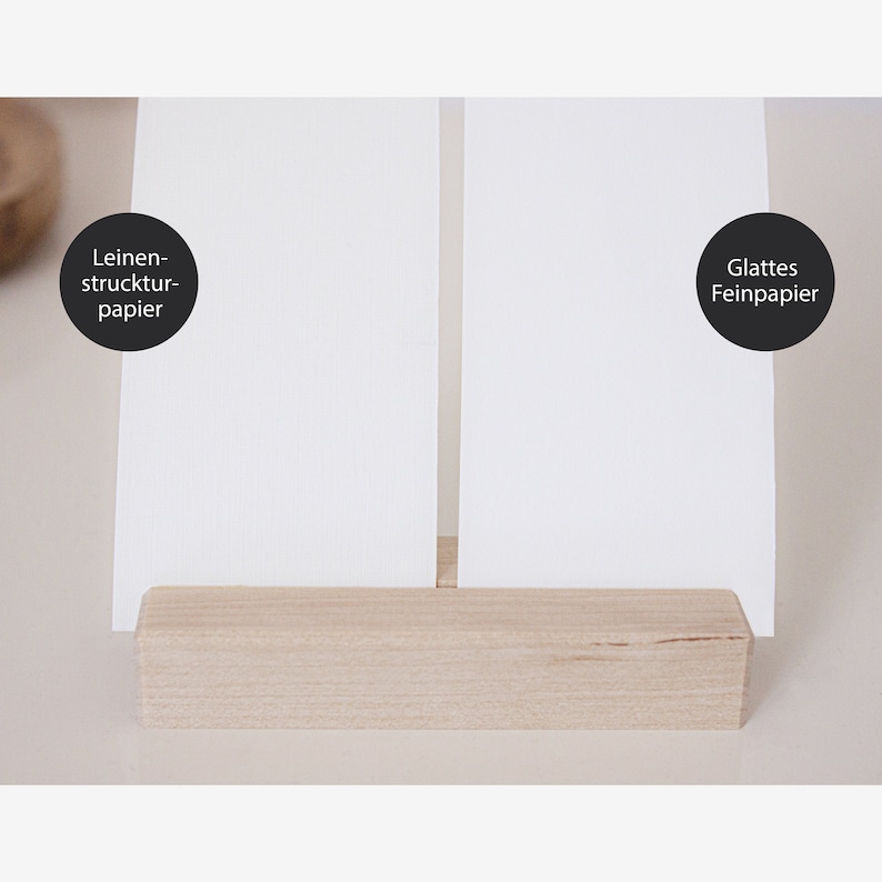 May include: Two white pieces of paper, one with a linen texture and the text "Leinen-struktur-papier" and the other with smooth texture and the text "Glattes Feinpapier", are displayed on a wooden stand.