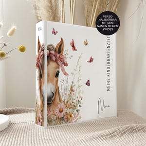 Könnte beinhalten: Ein weißer Ordner mit einer braunen Pferdeillustration, die einen rosa Blumenkranz trägt, umgeben von kleinen Schmetterlingen und Wildblumen. Der Text lautet "MEINE KINDERGARTENZEIT" und "Nina" in schwarzer Schrift. Ein schwarzer Aufkleber besagt "PERSONALISIERBAR MIT DEM NAMEN DEINES KINDES".