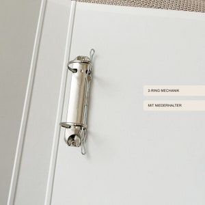 May include: A white binder with a silver 2-ring mechanism and a silver metal clip. The binder has the text "2-RING MECHANIK" and "MIT NIEDERHALTER" printed on it.