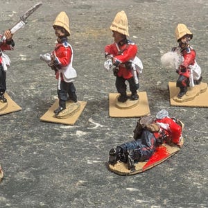 Puede incluir: Un conjunto de 10 soldados de juguete pintados, que representan tropas coloniales británicas con uniformes rojos y fajas blancas. Los soldados están en varias posturas, incluyendo de pie, de rodillas y tumbados. Algunos soldados sostienen rifles, mientras que otros sostienen bayonetas. Todos los soldados llevan cascos de pith y están sobre pequeñas bases marrones.