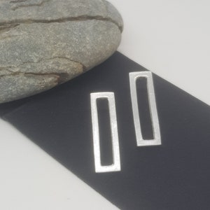 May include: Two silver rectangular earrings with a brushed metal finish. The earrings are displayed on a black surface, with a grey rock in the background. The earrings are a simple, modern design.