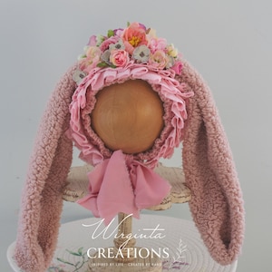 May include: A pink bunny bonnet with long, textured ears and a ruffled brim. The bonnet is adorned with a cluster of pastel-colored flowers and a matching pink ribbon. The bonnet is displayed on a wooden head form.