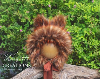 May include: A brown and orange faux fur bonnet with pointed ears and a rust-colored ribbon. The bonnet is set against a backdrop of green foliage and a wooden surface. The text "Virginia Creations" is visible.