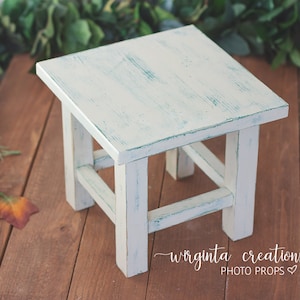 May include: White wooden stool with a distressed, light green paint finish. The stool has a square top and four legs with a simple, rustic design.