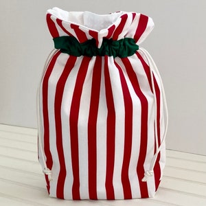 May include: A red and white striped drawstring bag with a green band at the top. The bag is made of fabric and has a white drawstring closure. The bag is designed to hold gifts or other items.